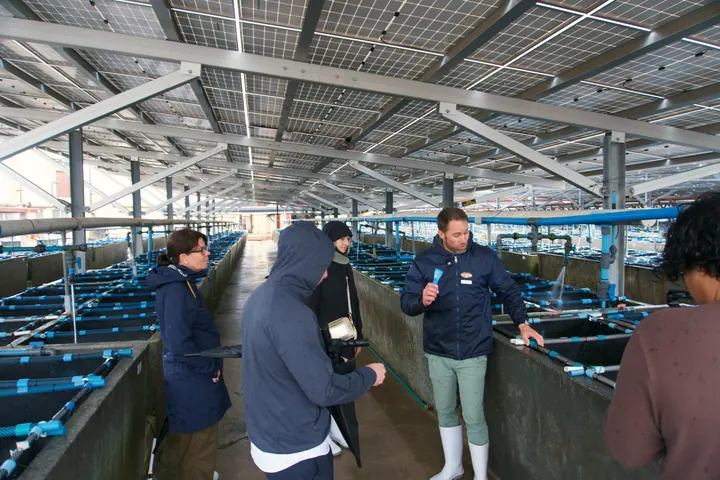 Jason and visitors talking beside rows of blue tanks inside an abalone farm