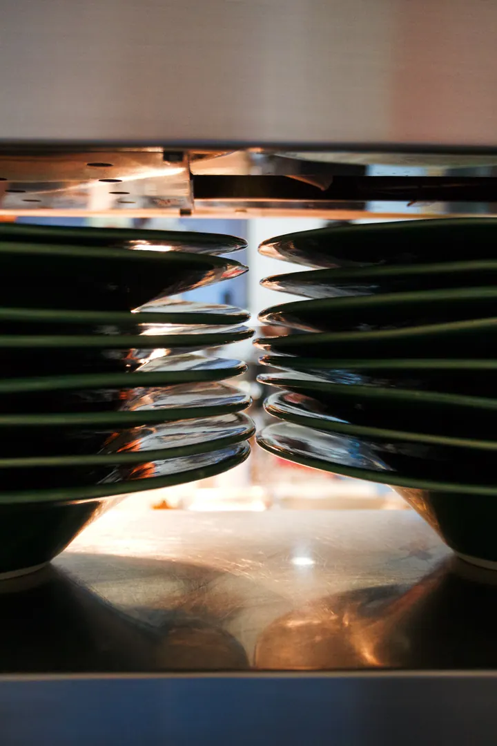 Stacks of dark plates warming under a kitchen heat lamp at Creation Wines