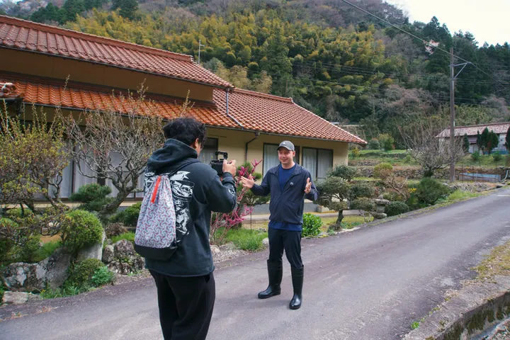 Gigi filming Jason beside a house on a road in the Japanese mountains