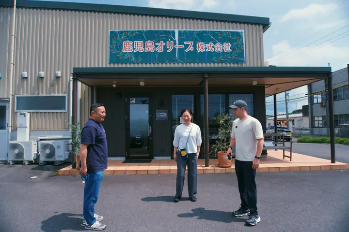 Three people talking outside a building marked Kagoshima Olive