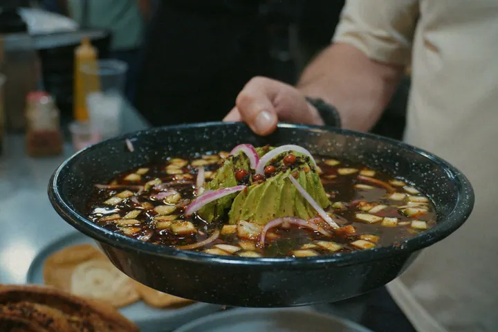 A hand holding a dark bowl of broth topped with avocado and onion