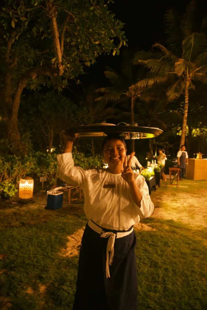 A smiling server carrying a tray overhead beneath palm trees at night